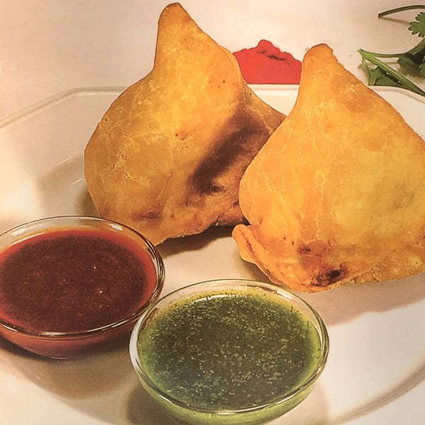 Two golden-brown samosas on a white plate with small bowls of red and green chutneys.