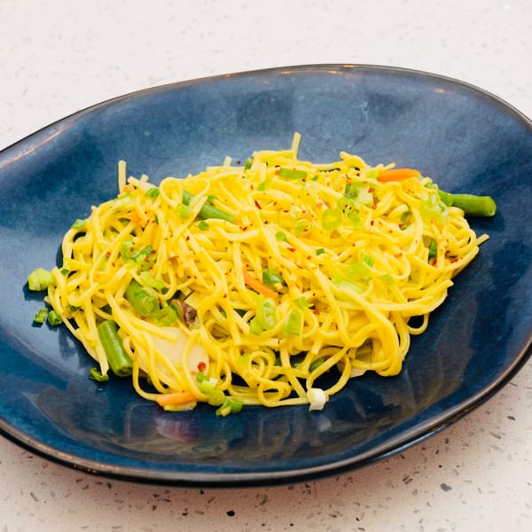 Yellow noodles with green onions, red pepper flakes, and other vegetables on a blue plate.