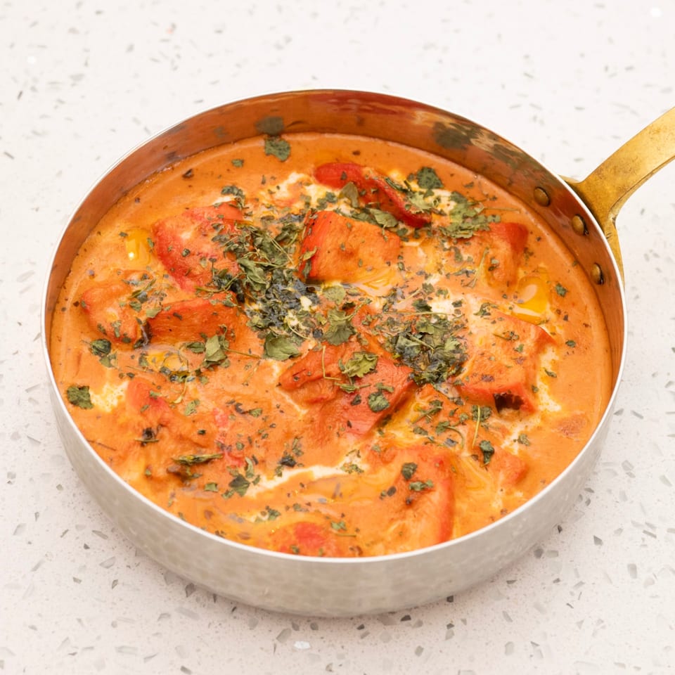 A copper pan filled with vibrant orange chicken tikka masala, garnished with fresh herbs.