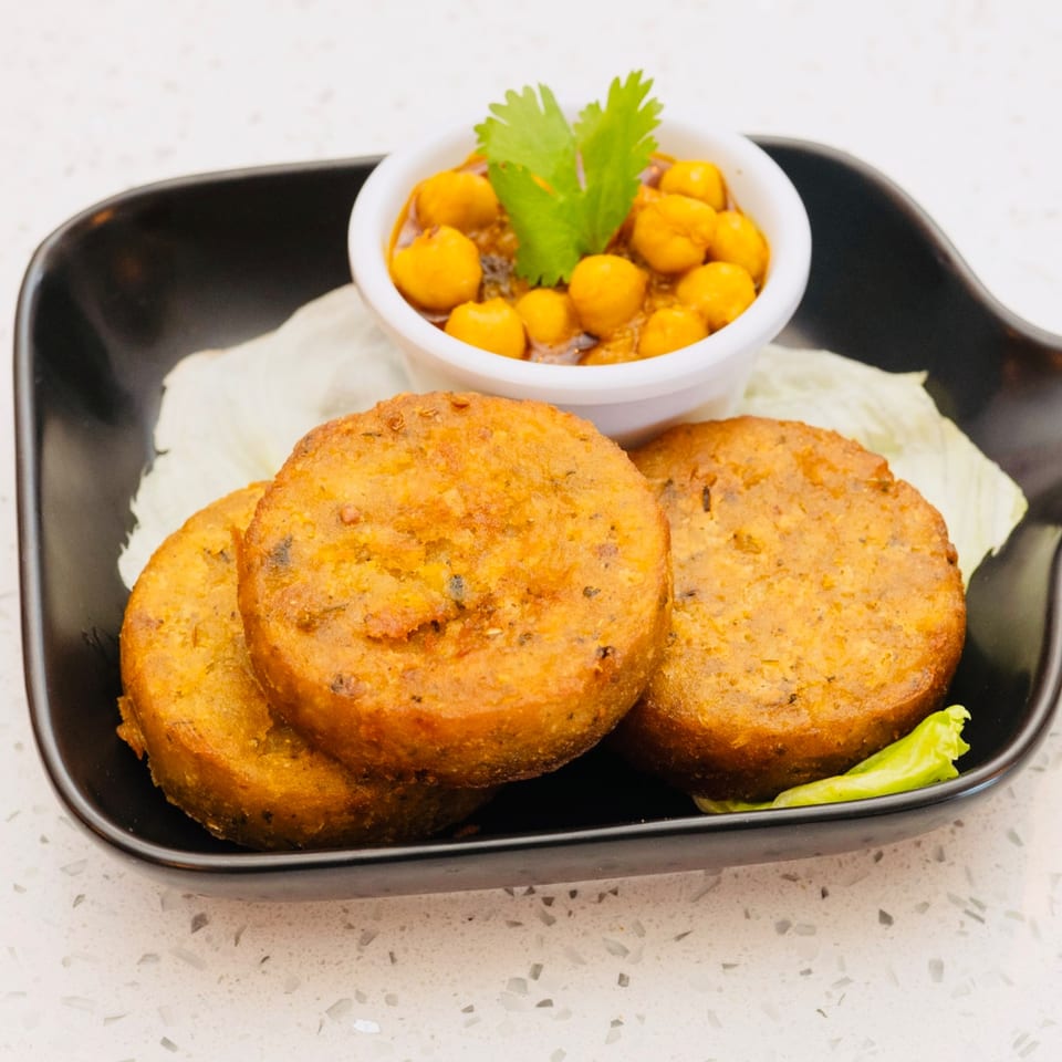 Three golden-brown potato patties with a side of chickpea curry and cilantro on a black plate.